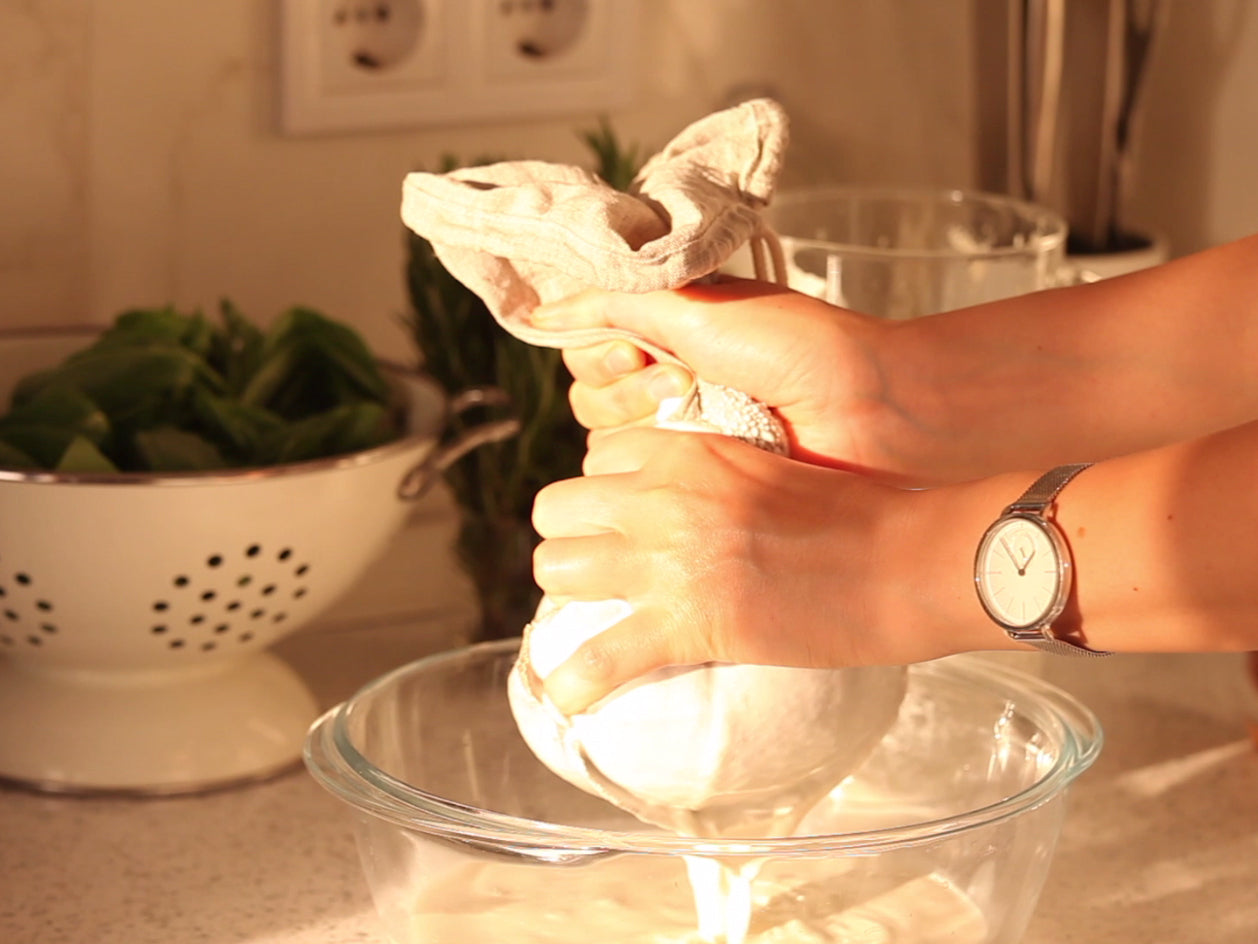 A Scene Where Almond Mash is Being Squeezed Through a Linen Nut Milk Bag and Almond Milk is Dripping Into a Bowl.