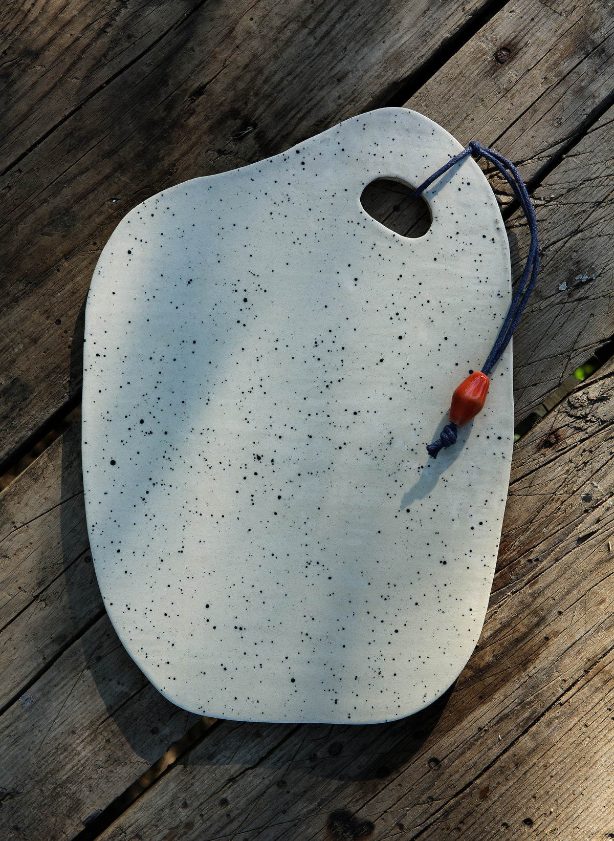 Speckled white ceramic cheese and charcuterie board with a blue cord and red ceramic bead on a rustic wooden table.