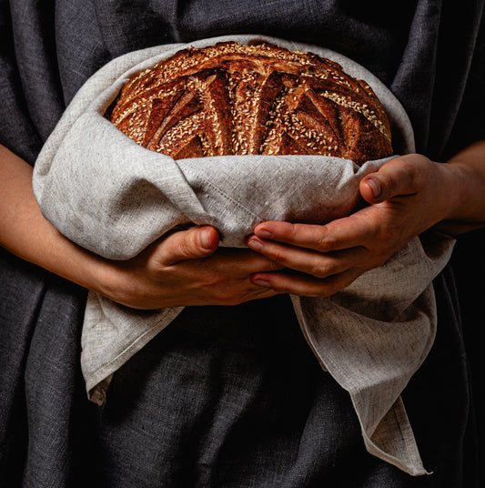 A pair of hands gently holds a round sourdough loaf with a golden crust, intricate scoring, and a dusting of sesame seeds. The bread rests inside an open linen bread bento bag, its soft folds framing the loaf like a nest.