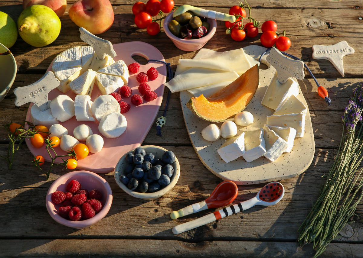 Assorted cheeses, fruits, and vegetables arranged on two ceramic cheese and charcuterie platters on a wooden table.