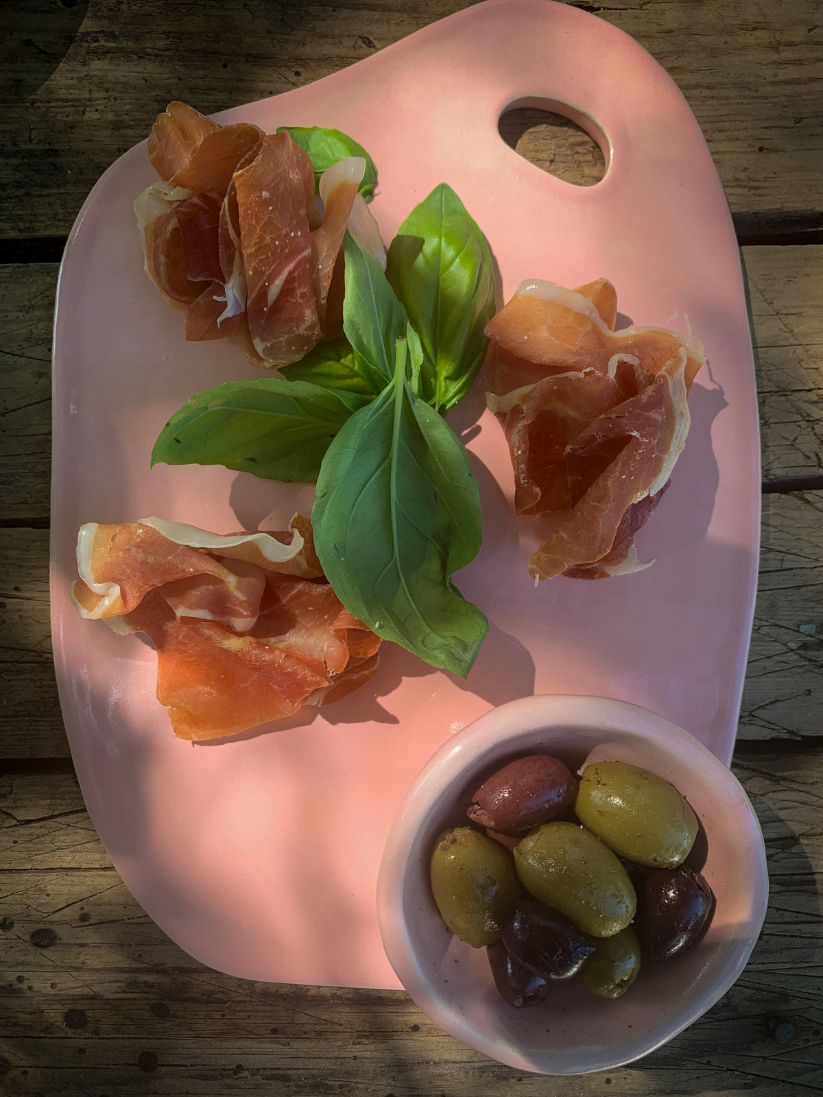 Pink ceramic cheese and charcuterie platter with prosciutto and basil, accompanied by olives in a small pink sauce bowl on a wooden surface.